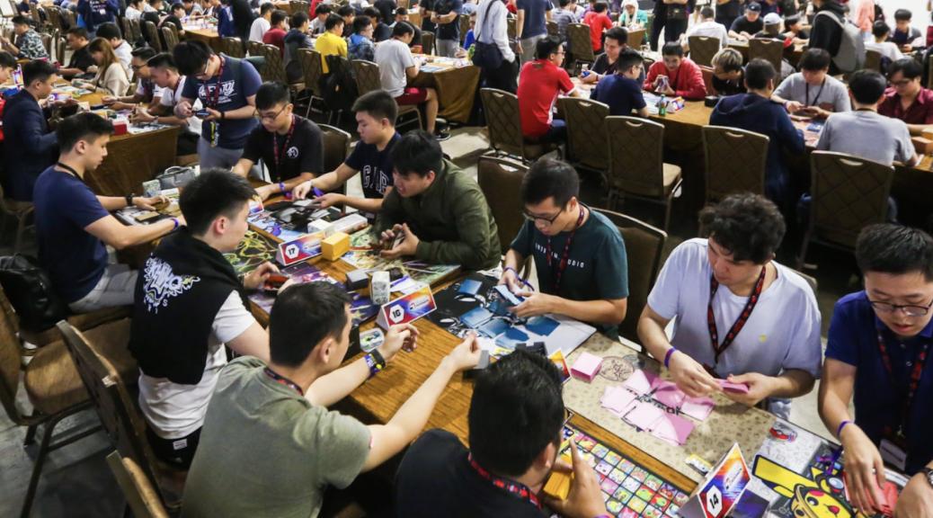 A packed tournament hall with rows of trainers battling at long tables