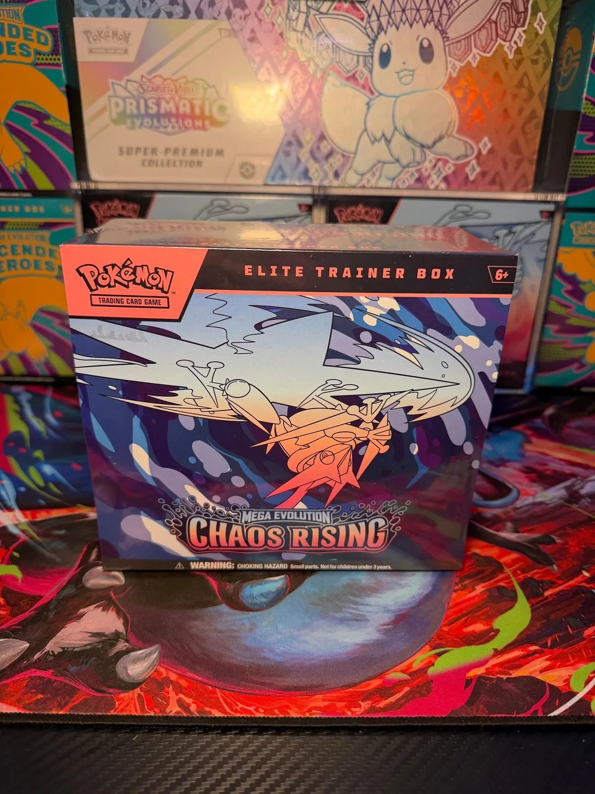 Pokémon Mega Evolution Chaos Rising Elite Trainer Box on a playmat with other Pokémon boxes behind it